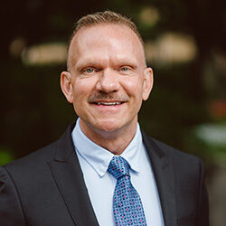 Ricky Bradley facing the camera for a headshot photo. Ricky is wearing a black coat, blue shirt, and a patterned blue tie.
