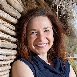 Outside portrait of Dr. Katherine Hayhoe. Dr. Hayhoe is standing by a rustic, wooden building, is wearing a blue shirt, and is smiling at the camera.