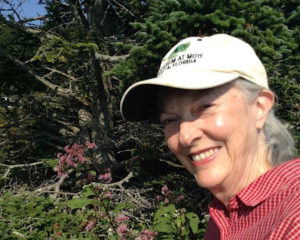 Norma Morrison wears a white ballcap and a red checkered button down, smiling in front of a blooming garden in Tennessee
