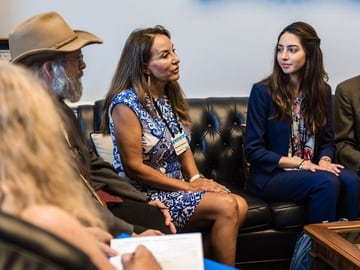 Three people engaged in a focused conversation in an congressional office. A man wearing a hat and a beard is listening intently to a woman in a blue and white dress. Next to the woman is a younger woman with long hair, wearing a blazer, who is also paying close attention to the discussion. They are seated on a black leather couch with lobbying handouts in hand.