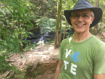 CCL volunteer Mark Taylor smiles at the camera while hiking a nature trail. He wears a green CCL shirt and a floppy black hat.