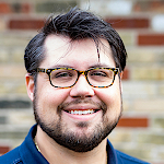 This headshot is of Alex Bozmoski, VP of Programs for DEPLOY/US. Alex sports a cheerful expression, sporting a pair of stylish glasses. He has short, dark hair and a beard. Dressed in a dark polo shirt, he appears casual yet neat. The background features a brick wall, suggesting an informal, possibly urban setting. 