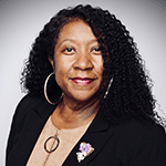 This is an image of Jennifer Burton is the Senior Energy Manager at Lowe’s, leading initiatives in energy management, efficiency, and renewable energy a professional woman with a friendly demeanor. She has curly black hair that falls to her shoulders and is wearing a black blazer. On her left lapel, she sports a delicate purple flower pin. Her attire is complemented with a simple, elegant necklace and hoop earrings. Jennifer is posed against a soft-focused background, which suggests a formal setting, possibly for a corporate headshot or professional profile.