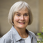 This is a headshot image of Mary Selkirk a retired collaborative environmental policy specialist. Mary has shoulder-length silver hair and a warm smile. She is wearing a light grey button-up shirt and appears outdoors, with blurred greenery in the background that suggests a calm, natural setting. 