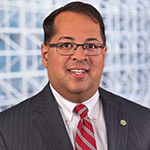 The image is of Neil Chatterjee the former Chairman of the Federal Energy Regulatory Commission. Neil is smiling with short black hair, wearing a dark suit, white shirt, and a red and white striped tie. He has a pin on his left lapel, indicating a potential affiliation with an organization or cause. The man appears to be in a professional setting, with an abstract patterned background that could be found in a corporate office.