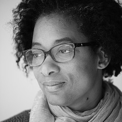 Artful, close-up, black and white headshot of Tina Johnson. Tina is wearing glasses and looking away from the camera, smiling slightly.