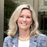 This headshot is of Zaurie Zimmerman, Citizens’ Climate Lobby’s Governing Board Chair. Zaurie has long blonde hair and a bright smile. She is wearing a tweed jacket and a yellow necklace that adds color to her business attire. The setting appears outdoors, with a reflective glass building in the background.