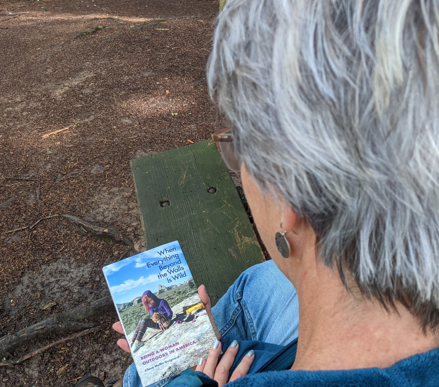Lilace Melin Guinard, wearing a blue shirt and blue jeans with grey hair and glasses, sits on a park bench and looks down at the cover of her memoir "When Everything Beyond the Walls is Wild: Being a Woman Outdoors in America."