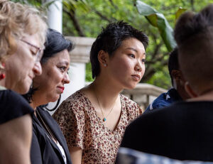 An Asian woman in a short sleeve floral dress sits to the right of a Hispanic woman in a dark blazer, and a white woman in a dark dress. They sit outdoors and listen to a member of their chapter speak.