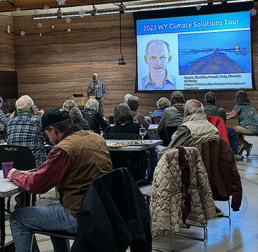 Bill Barron gives a presentation to his audience will touring through Wyoming. He stands on stage giving a presentation. In the foreground are attendees, including a man in a tan vest, a red shirt, and blue jeans.