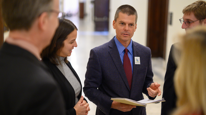 A middle-aged white man wearing a gray suit, blue shirt, and red tie stands in a hallway with a group of other volunteer climate advocates before a meeting with Congress. They look at him as he speaks, gesturing to a notebook in his hand.