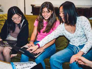 Three young female CCL volunteers sit closely together on a couch, engaged and smiling as they look at a laptop screen. The woman on the left, wearing glasses, is typing, while the one in the middle, in a pink shirt, points at the screen, and the one on the right, in a floral top, watches with interest. They are working together on climate solutions.