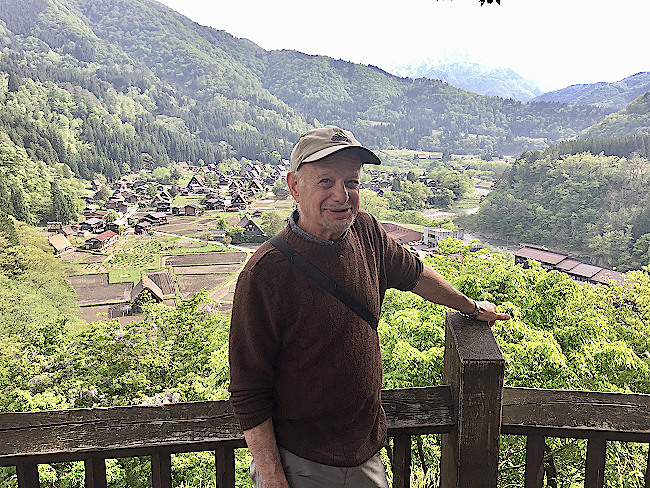 Donor Jim Lilienthal stands on a deck overlooking a lush green valley and foreign village. He wears a light colored hat and a brown sweater.