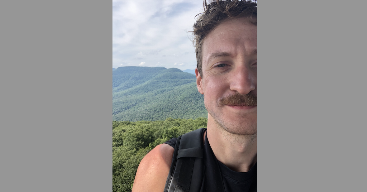 A selfie of Eric Dean Wilson, a smiling white man with a mustache, on a hike. There are tree-covered mountains in the background and white clouds in the sky.