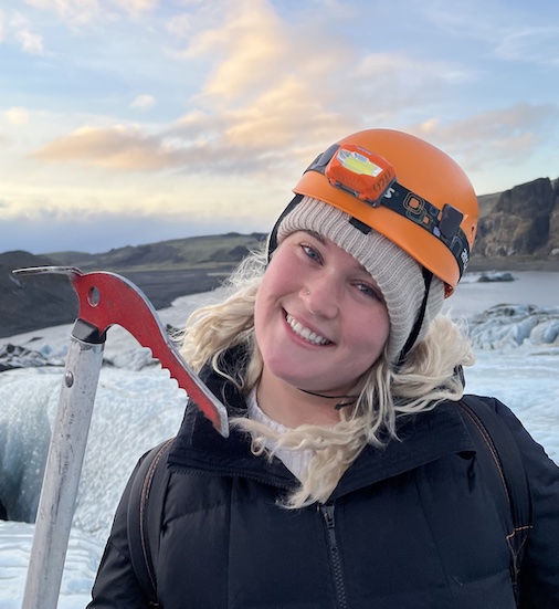 A white woman poses in an icy landscape, smiling, wearing a warm hat, helmet, and headlamp.