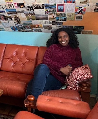 A black woman sits on an orange couch, smiling. The ceiling above her has a collage of photos and postcards.