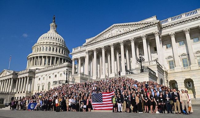 Large group of people stands on the steps of Capitol Hill holding an American flag. The Capitol dome is in the background.