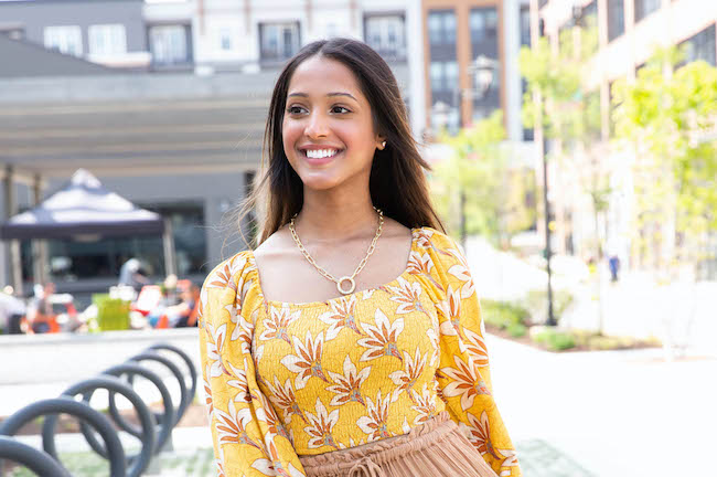 A woman stands on an urban street with a bike rack, buildings, and trees behind her. She is smiling. She has brown skin, long dark hair, and is wearing a yellow floral dress.