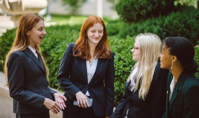 Four young women stand in a group, smiling at each other, wearing professional clothing