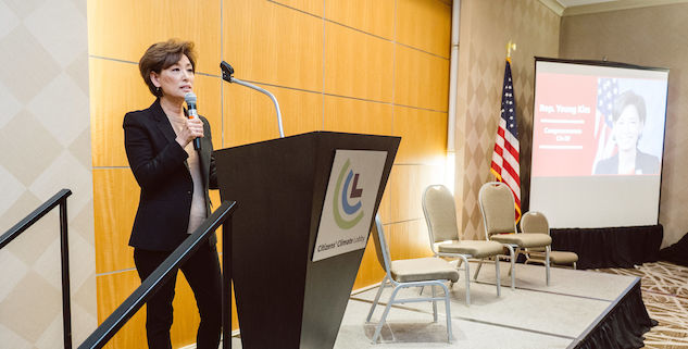 A Congresswoman stands behind a podium with a microphone. The stage next to her has three empty chairs, and a large screen showing her name and photo further off to the right. 