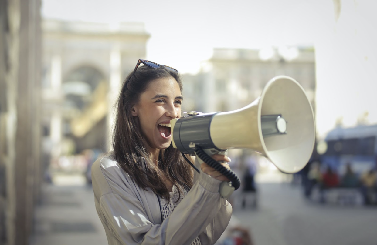 A white woman with long brown hair shouts cheerfully into a megaphone. The background is blurry but appears to be a city street.