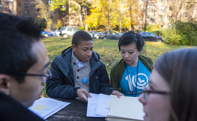 A young Black man and an Asian woman look at a stack of papers on a table. They are bundled up outside on a cold fall day.