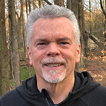 This is a headshot image of Jerry Hinkle, a leader with Citizens’ Climate Lobby since 2011. Jerry is wearing a black zip-up jacket and appears to be outdoors, with trees and autumn leaves in the background, indicating a natural setting. His casual attire and the outdoor environment give the impression of a relaxed and friendly individual.