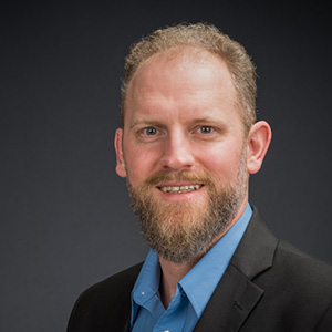 This headshot is of Professor Ross Astoria, who teaches law and policy at the University of Wisconsin, Parkside. Ross has a short, neatly trimmed beard and hair. He is wearing a dark suit jacket over a blue collared shirt. His expression is pleasant and confident, and he has a slight smile. The background is a simple, dark gradient, which provides a professional look suitable for business or academic purposes. 