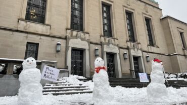 Three snowmen holding climate change protest signs stand in front of an official looking city building