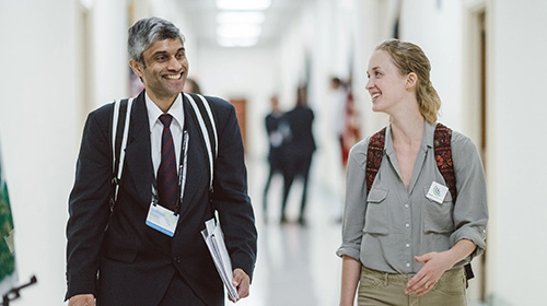Two CCL volunteers, a man and a woman, walking and conversing in a corridor on Capitol Hill. The man to the left is dressed in a dark suit, tie, and a lanyard with an ID badge, carrying papers under his arm. The woman to the right is casually dressed in a beige shirt, with a backpack over her shoulder and a lanyard with an ID badge. They both wear cheerful expressions and appear engaged in a pleasant conversation.