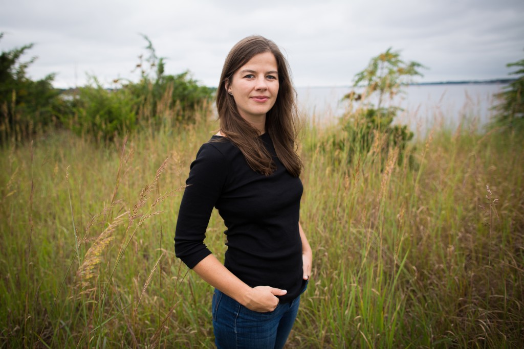 A white woman in a black shirt stands in a marsh, smiling at the camera