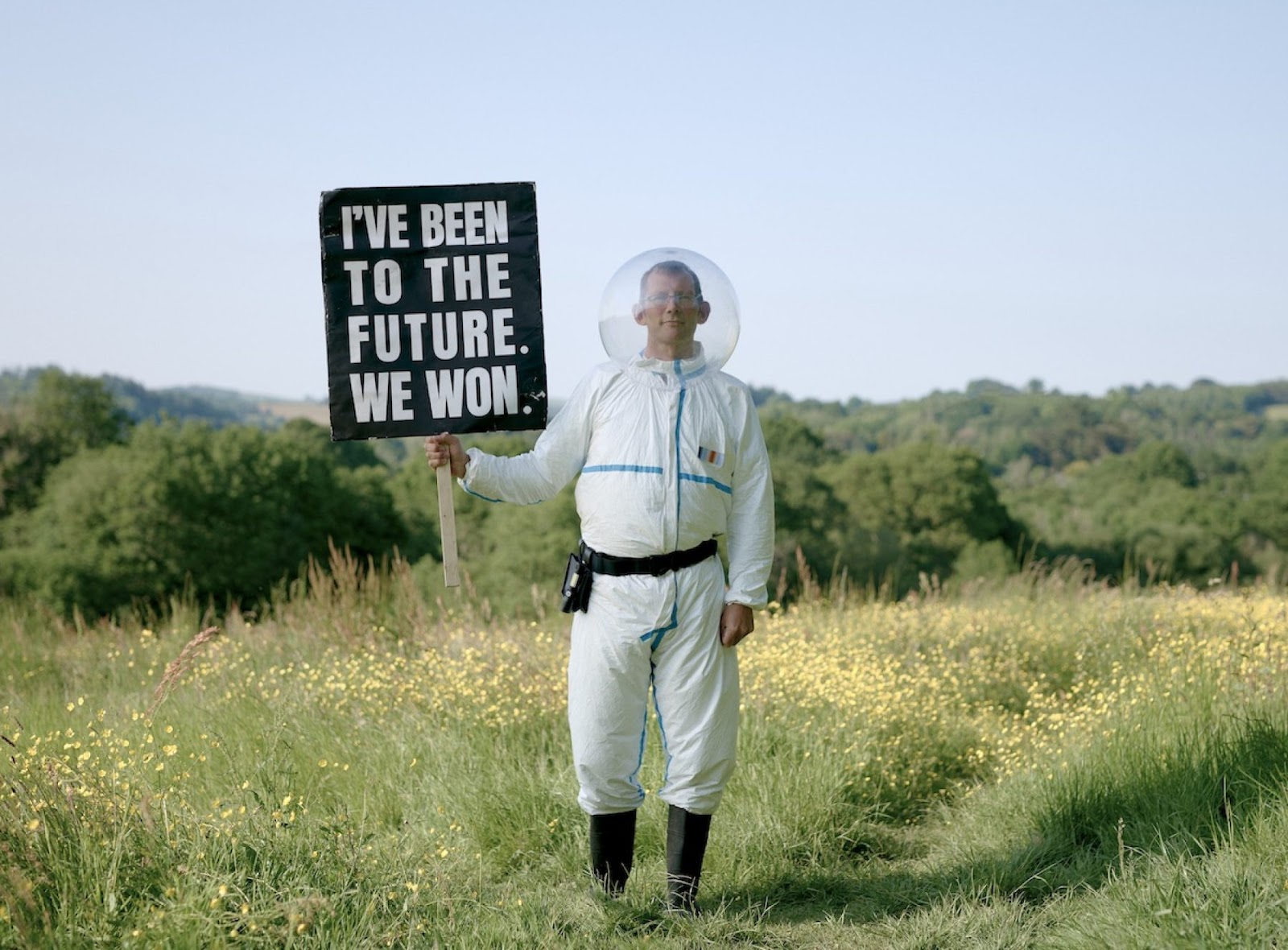 Rob Hopkins stands in a field wearing a futuristic suit with a bubble around his head, holding a sign that reads I've Been to the Future, We Won.