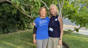 Two older white women stand together in front of a tree. One has her arm around the other, and they are both smiling at the camera.