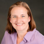 Headshot of Rachel Kerestes, a Caucasian woman with shoulder-length light brown hair, smiling warmly at the camera. She is wearing a light purple blouse and is photographed against a soft-focused, dark gray background.