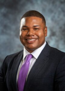 A smiling Black man wears a dark suit, white shirt, and purple tile.