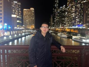 An Asian man leans agains the railing of a bridge at night with a city skyline in the background.