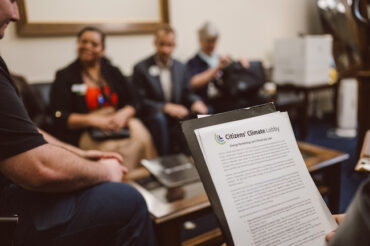 Women and men in suits sit around a coffee table in an office waiting room. They are blurry in the background. In the foreground is a sheet of paper with typed writing on it, held up as if someone is reading from it.