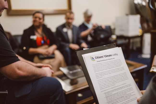 Women and men in suits sit around a coffee table in an office waiting room. They are blurry in the background. In the foreground is a sheet of paper with typed writing on it, held up as if someone is reading from it.