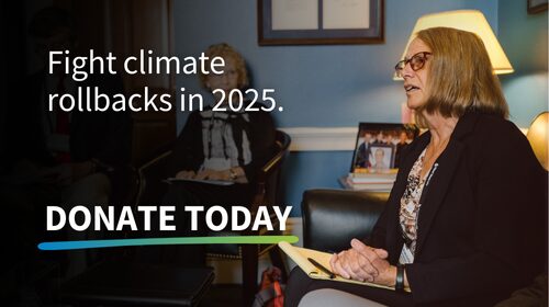 An image of a woman sitting in a professional setting, holding a notepad and speaking. The text overlay reads: 'Fight climate rollbacks in 2025. DONATE TODAY,' with a multicolored underline beneath 'DONATE TODAY.' The scene includes warm lighting, a lamp, and a framed photo in the background, conveying a call to action for climate advocacy.