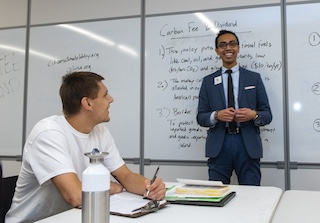Two men in an office, one at a desk with a notebook and one standing next to a whiteboard.