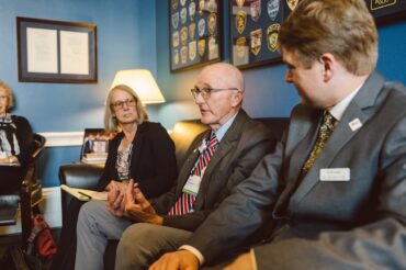Two men and one woman sit on a couch in a congressional office dressed in professional attire. The man seated in the middle is speaking.