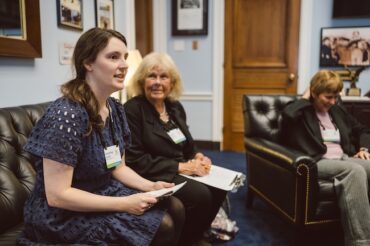 Three women sit in a congressional office. One is speaking to someone who sits off camera