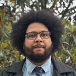 A man with a full beard, wearing glasses, a patterned dress shirt, a patterned tie, and a dark overcoat, standing in front of a background of leafy trees.