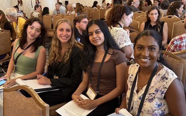 Four young women of different ethnicities are sitting in a row of conference seating. They are wearing nametag lanyards, holding folders with papers on them, and smiling at the viewer