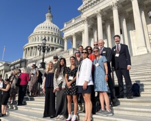 A group of people (various ages, genders and ethnicities) stand on steps with the U.S. Capitol dome in the background
