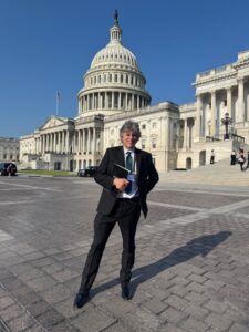 A white man with gray hair wears a black suit, a lanyard around his neck, and holds a folder of materials. The U.S. Capitol is in the background.