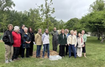 A group of people stand shoulder to shoulder in a grassy area, surrounding a newly planted tree. Mature trees and blue sky are in the background.
