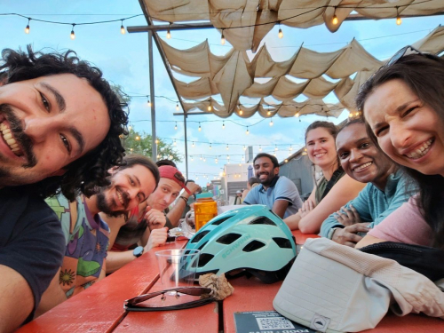 Smiling people in their 30s sit on either side of a picnic table and lean into the frame of the photo, against the background of a sunny sky. A bike helmet and sunglasses are on the table.