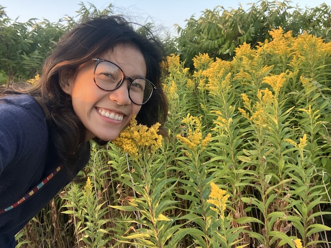 A smiling woman with light brown skin and brown hair, wearing glasses, leans toward bright yellow flowers