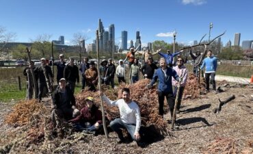 A group of people stand outside smiling and holding branches, with a sunny city skyline behind them